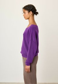 Lilla strikket sweater med bred hals, sænkede skuldre og ribbede ærmer, parret med brune bukser, vist i profil mod en lys baggrund.