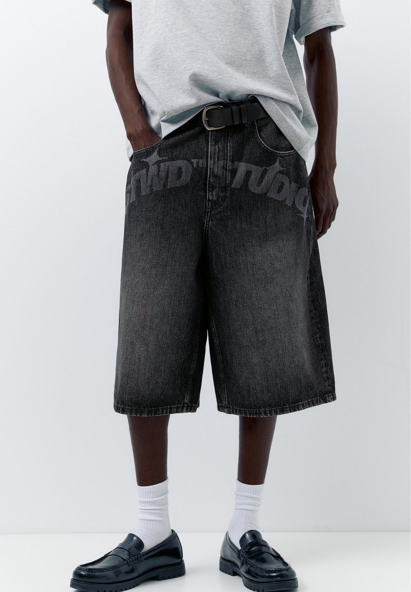 Personne portant un short en jean noir délavé oversize avec une ceinture noire, un t-shirt gris, des chaussettes blanches et des mocassins noirs, les mains dans les poches.