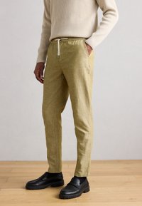 Ανοιχτό κίτρινο, υφασμάτινο παντελόνι με ελαστική μέση και κορδόνι. Διαθέτει πλαϊνές τσέπες και σχεδιασμό με στενή γραμμή στα πόδια. Μαύρα παπούτσια συμπληρώνουν το look.