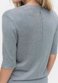 Kvinna som bär en ljusgrå stickad topp med ärmar som når till armbågen och en vertikal söm med en liten metallring på baksidan.