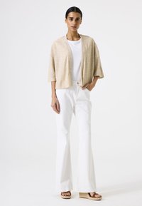 Beige gestrickte Strickjacke mit weiten Ärmeln, die über einem weißen Hemd getragen wird. Kombiniert mit weißen Schlagjeans und braunen Keilsandaletten, steht vor einem schlichten Hintergrund.