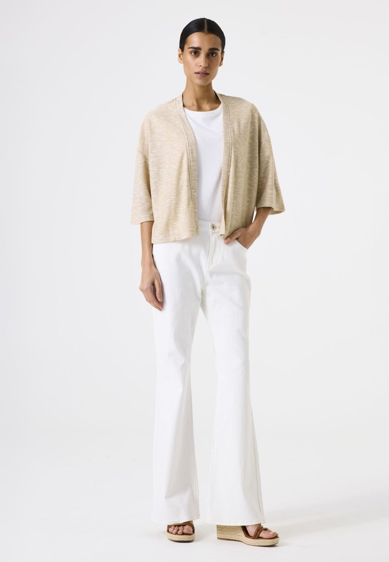 Beige gestrickte Strickjacke mit weiten Ärmeln, die über einem weißen Hemd getragen wird. Kombiniert mit weißen Schlagjeans und braunen Keilsandaletten, steht vor einem schlichten Hintergrund.