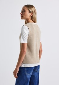 Top sin mangas beige de punto sobre una camisa blanca, con textura de canalé y un escote redondo. Combinado con pantalones de mezclilla azules.