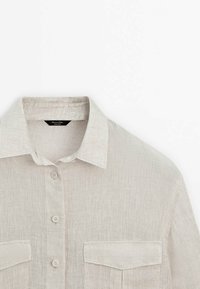 Chemise en lin beige avec col, patte de boutonnage sur le devant et deux poches poitrine, présentée sur un fond blanc.