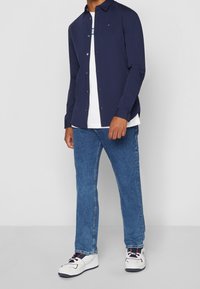 Man som bär en marinblå skjorta med knappar över en vit grafisk t-shirt, blå jeans och vita sneakers med marinblå och röda detaljer, stående på en grå golv.