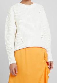 Maglione bianco lavorato a maglia con scollo rotondo, texture a coste, spalle scese e spacchi laterali, abbinato a una gonna arancione fluente.