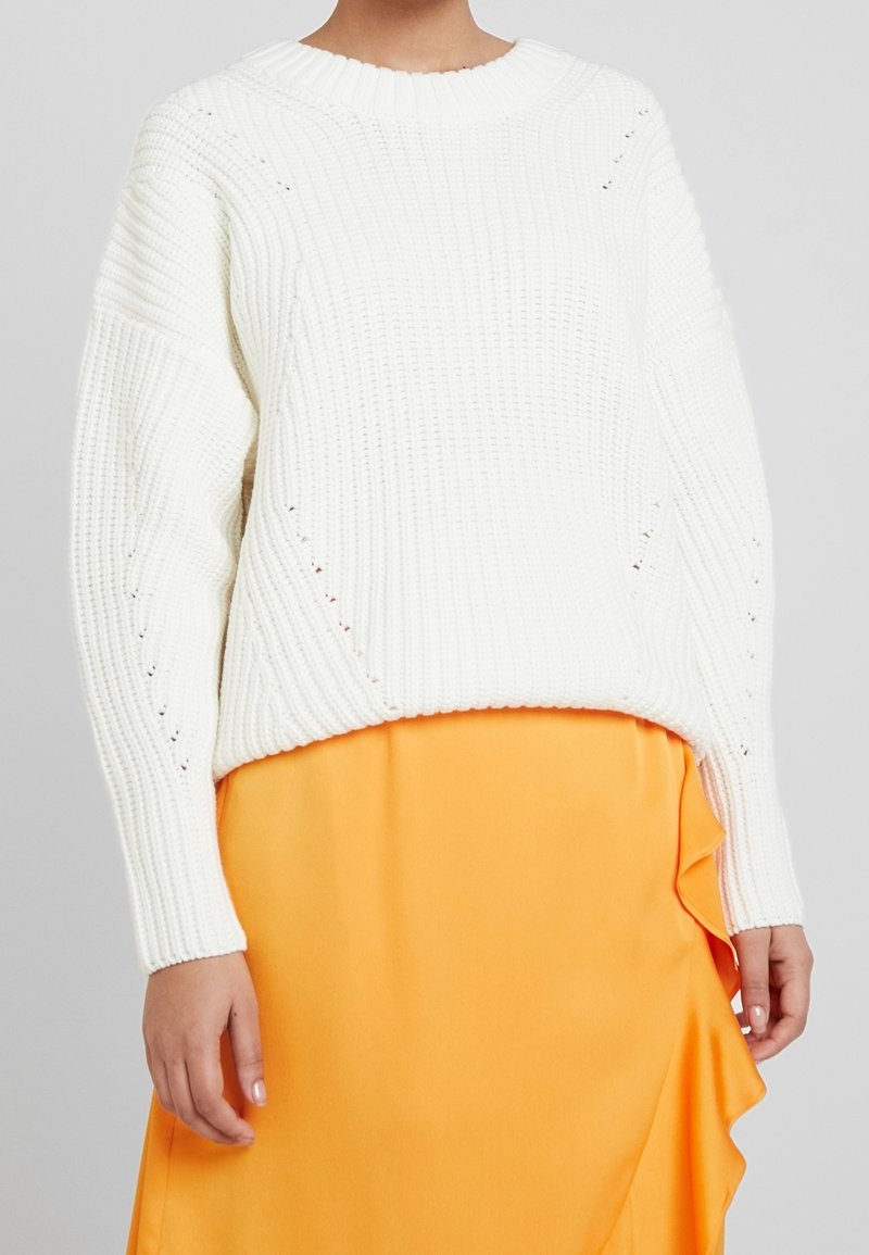 Maglione bianco lavorato a maglia con scollo rotondo, texture a coste, spalle scese e spacchi laterali, abbinato a una gonna arancione fluente.