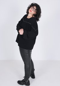 Zwart oversized sweater met studs, gecombineerd met glanzende zwarte nep leren leggings en veterschoenen. Krullend haar en een bril.