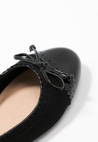 Chaussure noire avec un embout en cuir verni, corps en daim, perforations décoratives et un nœud attaché à l'avant. Conception plate.