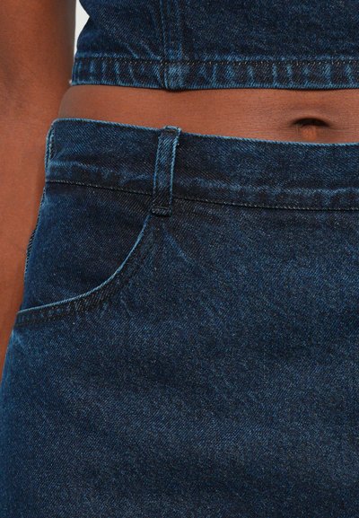 Tumma sininen denim-kangas, jossa on lähikuva istuvasta vyötäröosasta ja taskusta. Ompeleen yksityiskohdat ovat näkyvissä, mikä parantaa tekstuuria.