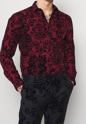 Homme portant une chemise rouge à manches longues avec un motif floral damassé noir et un pantalon noir avec un imprimé floral subtil, mains dans les poches.