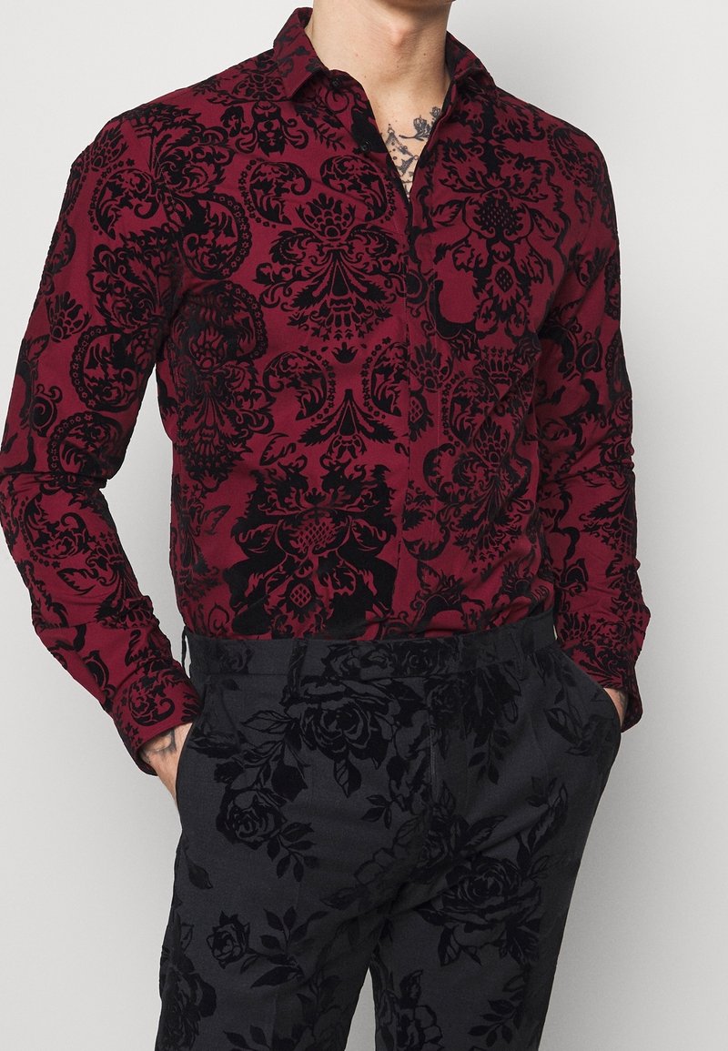 Homme portant une chemise rouge à manches longues avec un motif floral damassé noir et un pantalon noir avec un imprimé floral subtil, mains dans les poches.