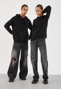 Svarte hettegensere med snøring og frontlommer, båret over slitte, oversized grå jeans. Jeansene har store rifter nær knærne. Svarte støvler.