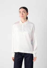 Blouse en satin blanc avec un col, un passepoil à boutons et des manches longues. Présente une broderie logo subtile sur le côté gauche. Associée à un jean foncé.