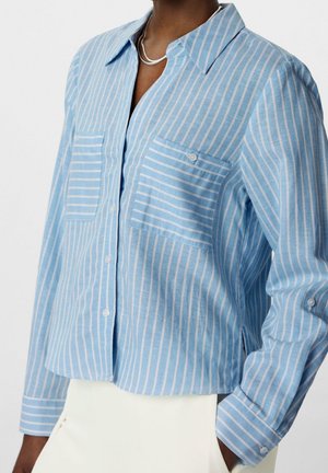 Blauw-wit gestreept overhemd met verticale strepen en knopen, met borstzakken, gedragen met witte broek en een zilveren kettinkje.