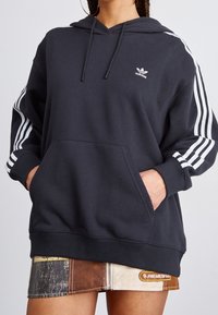 Sudadera gris oscura hecha de tela suave, con un bolsillo frontal, capucha con cordón y acentos de tres rayas blancas en las mangas.