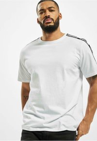 White cotton t-shirt with short sleeves, round neckline, and black accent stripes on shoulders featuring logo text. Smooth texture, relaxed fit.