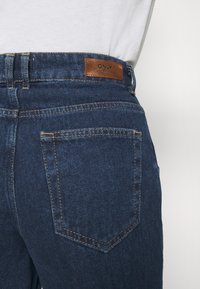 Tmavě modré džínové kalhoty mají koženou cedulku na pasu, dvakrát prošité švy a jedinou zadní kapsu s diagonálním střihem.