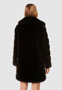 Cappotto di pelliccia nera con una finitura morbida e testurizzata, strisce orizzontali, colletto ampio e maniche lunghe, che arriva a metà coscia.