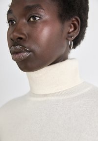 Κρέμα ζιβάγκο μπλούζα με ριμπ ύφανση, που διαθέτει ψηλό γιακά. Το μοντέλο φορά μικρές σκουλαρίκια κρίκους. Λείο δέρμα και γυαλιστερά χείλη.