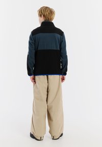 Fleece jack in zwart en teal met een hoge kraag, gecombineerd met beige, losse broek. De jack heeft een blauwe accent bij de zoom.