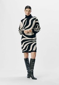 Robe en tricot avec un col haut dans un motif zèbre noir et crème ; manches longues et bouffantes ; assortie à des bottes noires à talons pointus arrivant au genou.
