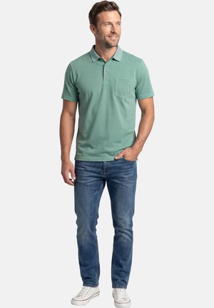 Homme portant un polo vert à manches courtes, un jean bleu et des baskets blanches, debout avec une main dans la poche, regardant vers la droite et souriant.