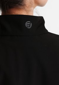 Persona con una chaqueta negra de alto cuello que muestra un pequeño logotipo circular gris en la parte posterior del cuello.