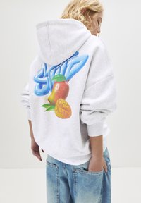 Giovane con felpa grigia chiara con testo in stile graffiti blu e grafica di mango, abbinata a jeans blu larghi, in piedi di lato.