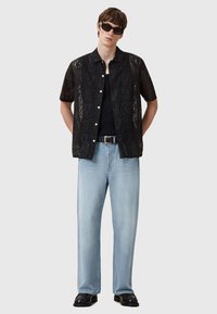 Camiseta negra de manga corta con un patrón similar al encaje, llevada sobre una parte superior negra, combinada con jeans rectos de color azul claro y zapatos negros. Gafas de sol negras.