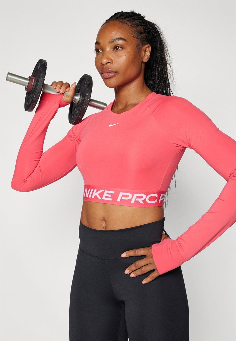 Długi rękaw, krótki top Nike Pro w jasnoróżowym kolorze z białym logo; połączony z czarnymi legginsami z wysokim stanem i trzymający hantle.