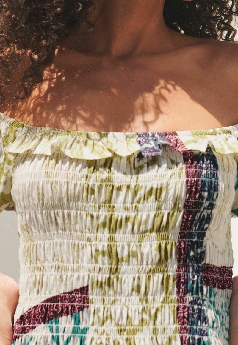 Top bustier à épaules dénudées avec un design smocké en blanc, vert et bordeaux, avec des bords à volants et un tissu à motif coloré.