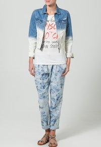 Denimjakke med gradient blå nuancer, parret med en hvid T-shirt med rød og sort tekst, og lyseblå tie-dye bukser. Brune snøre sandaler.