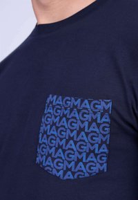 T-shirt en coton bleu marine avec une poche à motifs comportant un texte "MAG" répété dans une teinte de bleu plus foncé. Design classique à col rond.