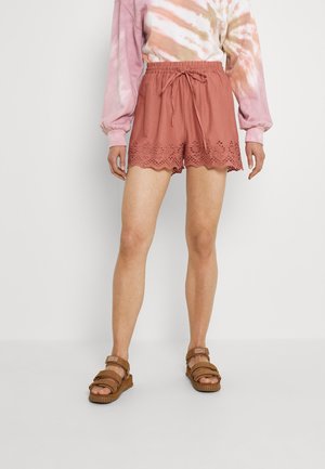 Person iført rustfarvede shorts med øjer og snørebånd i taljen, en pink og beige tie-dye sweatshirt og brune sandaler.