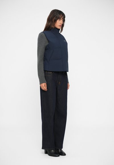 Námornícky modrý vypchaný vest s vysokým golierom, dva predné vrecká a šedý dlhý rukáv top pod ním. Model nosí tmavé denimu široké nohavice a čierne čižmy.