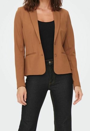 Mujer con blazer marrón ajustado sobre una camiseta negra y vaqueros oscuros, de pie con los brazos relajados frente a un fondo liso.