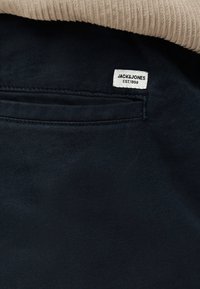 Τσέπη σκούρου υφάσματος με μια μικρή λευκή ετικέτα που δείχνει "JACK&JONES EST. 1990", μερικώς καλυμμένη από μπεζ ριγωτό ύφασμα.