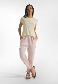 Żółty t-shirt z krótkim rękawem z nadrukiem w formie szkicu, zestawiony z różowymi lnianymi spodniami z elastyczną talią i bocznymi kieszeniami. Brązowe sandały.