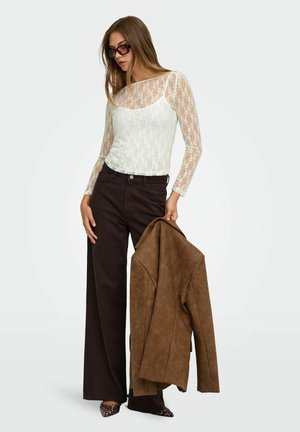 Jeune femme portant des lunettes de soleil, un top en dentelle blanche, un pantalon large foncé, des chaussures à motif léopard, tenant une veste marron dans sa main droite.