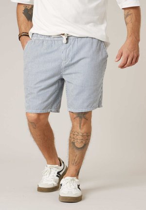 Homme portant des baskets blanches et un short à cordon rayé bleu et blanc, debout les mains partiellement dans les poches.