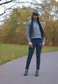 Sportowy strój składający się z szarej bluzy, czarnych spodni i czarnych sneakersów. Długie warkocze, stojąc na ścieżce z zielenią w tle.
