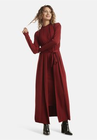 Long burgundy dress with long sleeves, a fitted bodice, and a tie belt at the waist. Smooth texture, floor-length design, and side slit details.