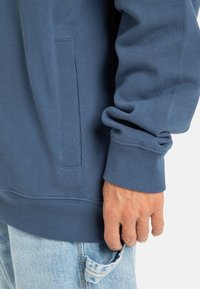 Sudadera azul hecha de tejido texturizado, con un bolsillo lateral y puños acanalados. El dobladillo también es acanalado para un ajuste ceñido.