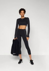 Schwarzes, langärmliges, bauchfreies Oberteil und hoch taillierte Leggings mit kontrastierenden weißen Seitenähten, kombiniert mit schwarzen Turnschuhen und einer schwarzen Jacke.