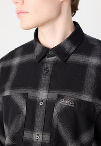 Černá a šedá kostkovaná flanelová košile s límečkem, obsahující kapsu na levé hrudi a vyšívané logo "BARBOUR INTERNATIONAL".