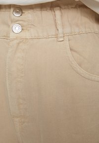 Zbliżenie na beżowe dżinsy, ukazujące dwa metalowe guziki, pętelkę na pasek oraz kształtowe przeszycia przedniej kieszeni.