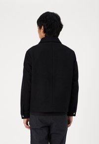 Veste matelassée noire avec col, manches longues et coupe droite. Comprend des boutons-pression sur les poignets et une texture de tissu lisse.