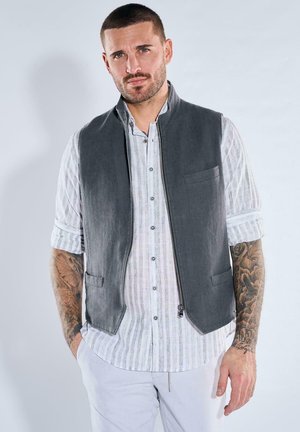 Man with short hair and tattoos wearing a rolled-up striped shirt, dark gray sleeveless vest, and light-colored pants, hands in pockets.