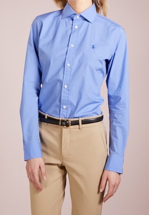 Persoon draagt een lichtblauw overhemd met knopen, ingestopt in beige broek met een zwarte riem, staand tegen een effen achtergrond.